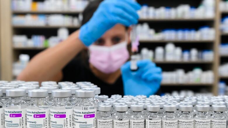 Small bottles of COVID vaccine are seen lined up inside a pharmacy, as a pharmacist in the background draws a dose while wearing protective gloves and a mask.