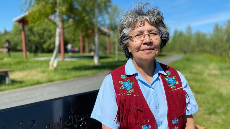 A woman in glasses and a beaded vest sits outside in the summer.