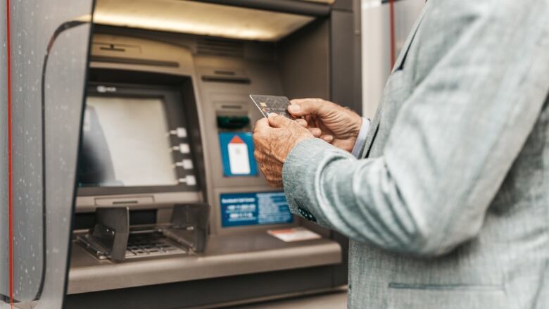 A older person with wrinkled hands, wearing a grey suit jacket, is shown holding a bank card in front of an ATM machine.