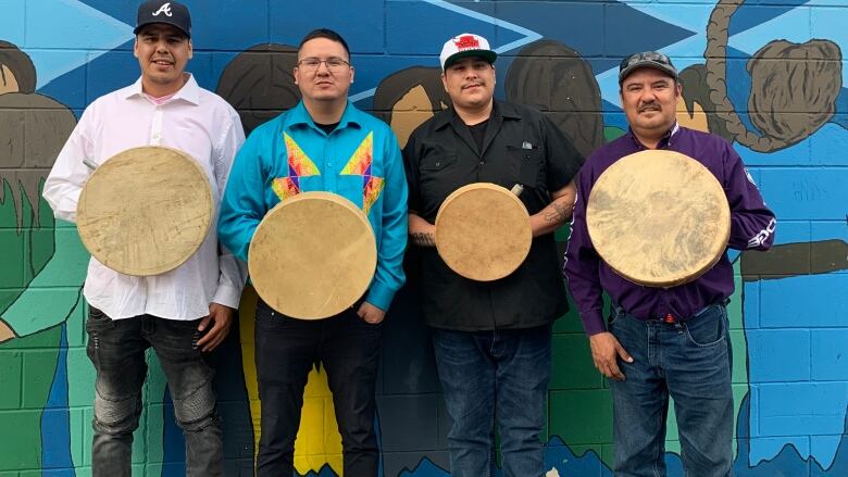 Four men with ball caps on stand against blue wall holding hand drums.