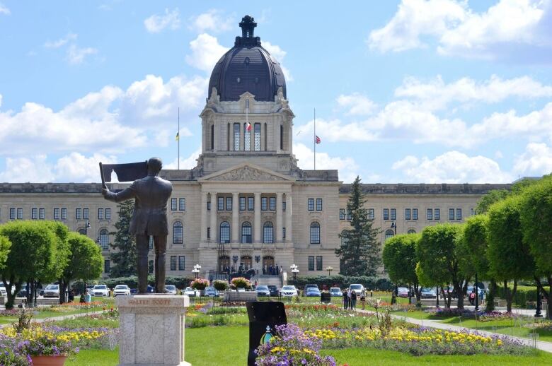 A blue sky with white puffy clouds hang above a government building with a tower. The building stands behind a parking lot filled with vehicles and a garden of flowers of red, purple and yellow. There's a statue erected in the garden. Trees line the edge of the green space.