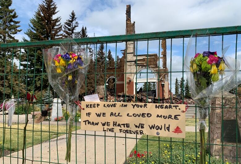 Two small bouquets of flowers and a handwritten note are attached to a green wire fence surrounding the burned remains of a church building. The note reads: Keep love in your heart. If we all loved more than we hate we would live forever.