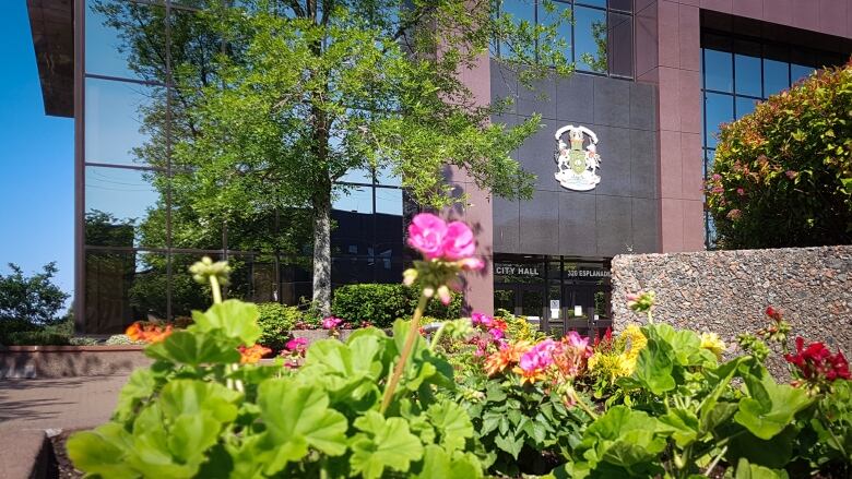 Pink and green flowers bloom in the sunshine in the foreground while a brown brick and blue glass building with a city hall sign and crest are in the background.