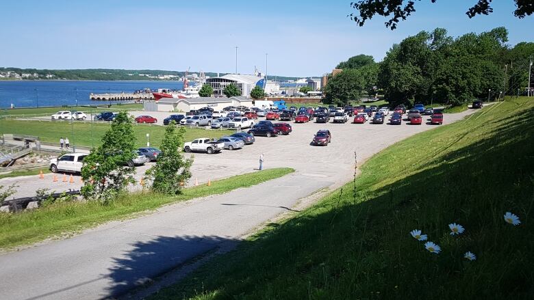 A gravel parking is shown on land surrounded by green grass next to a waterfront harbour.