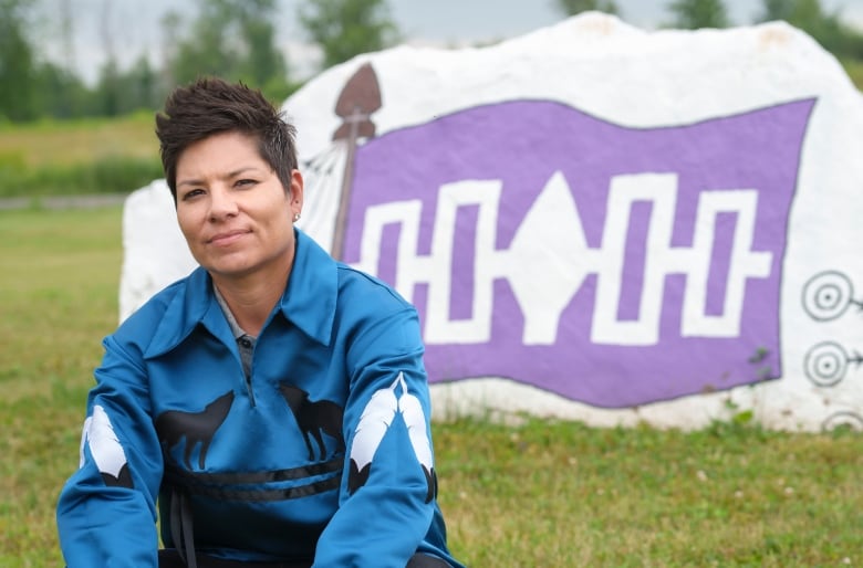 A woman wearing a ribbon shirt sits on the grass in front of a large rock painted with the Haudenosaunee Confederacy flag.