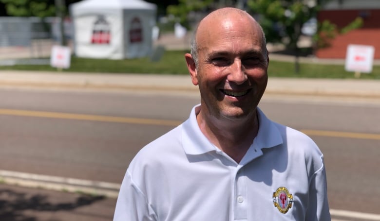 A man with short grey hair smiles at the camera, standing outdoors next to a road. He's wearing a white polo shirt.