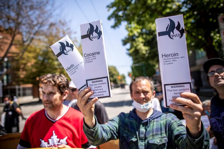 A man wearing a mask around his chin holds up three boxes, styled like cigarette cartons. They are marked 'Meth', 'Cocaine' and 'Heroin'. The man is surrounded by other people.