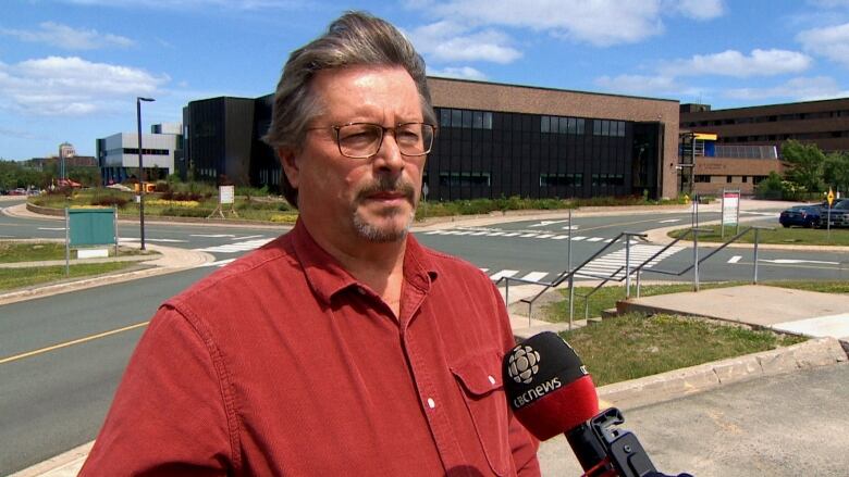 A man is standing outside a large building. He has glasses and the wind is blowing his hair. 