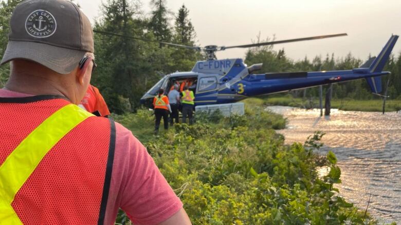 A man wearing a reflective yellow and orange vest stands toward a search and rescue helicopter in the middle of a forest near water.