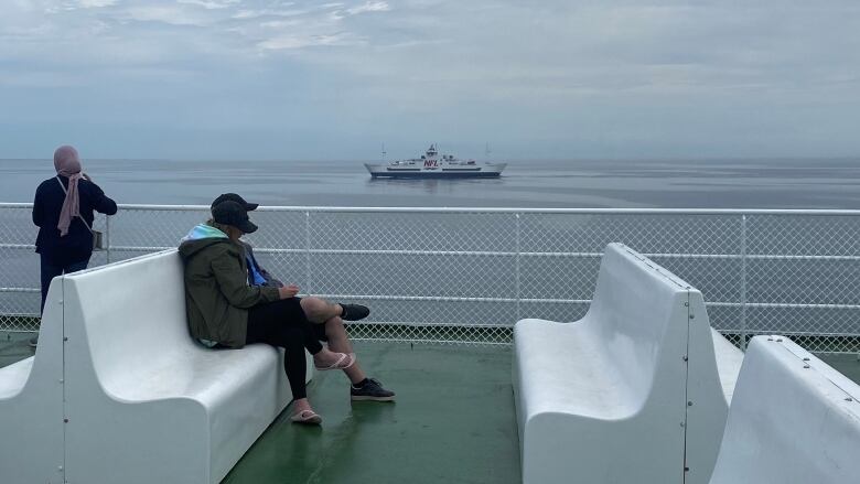 Three passengers stand on the green-painted deck of a passenger ferry in the middle of a stretch of blue ocean.