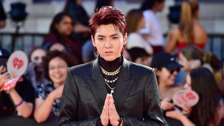 A man wearing a black leather jacket with a black turtleneck, with gold chains around his neck, stands with his hands pressed together in front of a crowd of people.