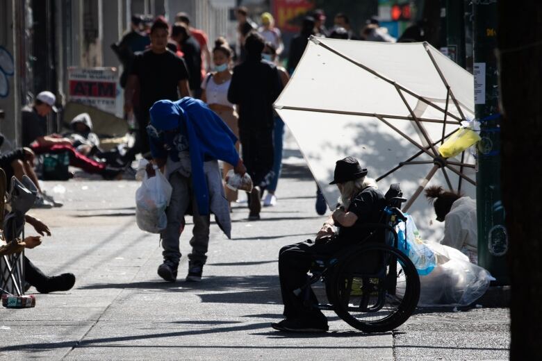 A large umbrella blocks the sun as people sit on the sidewalk.