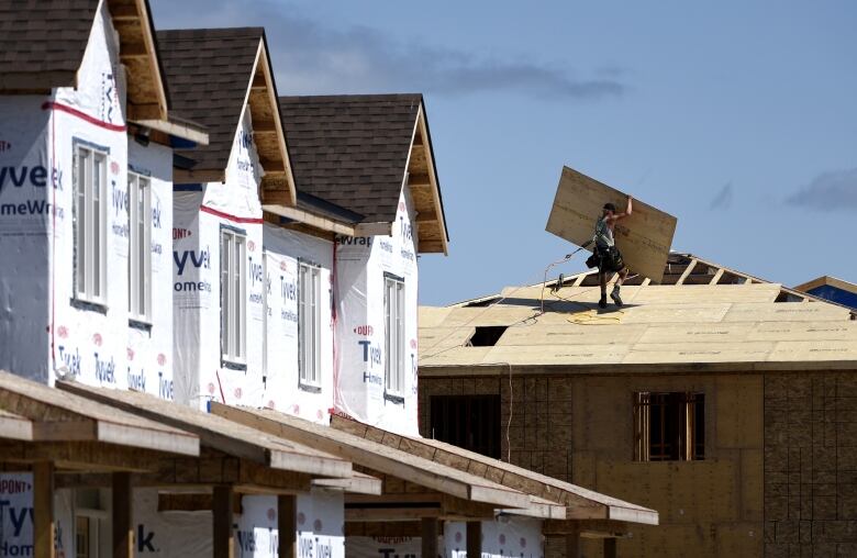 A man carries a piece of plywood across the roof of a home under construction