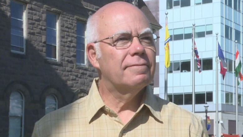 A man with glasses and a yellow shirt listens to a reporter's question near an outdoor rally. 