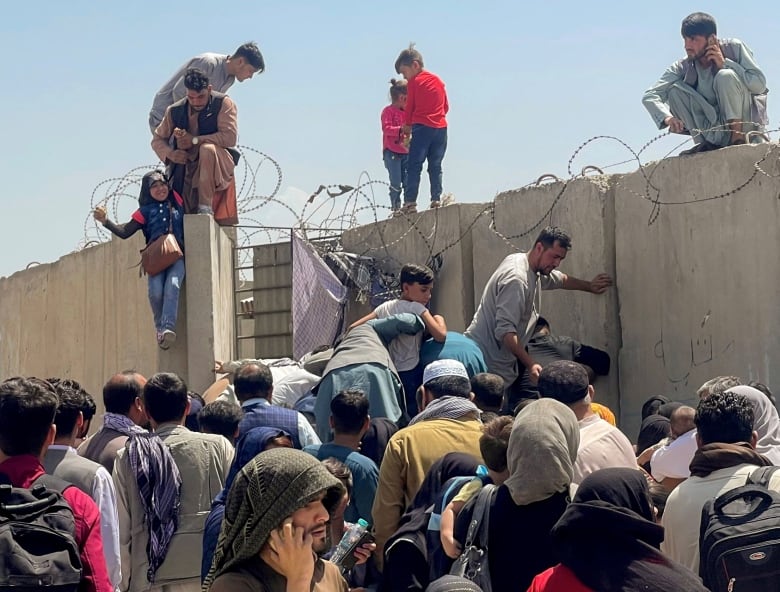 Men, women and children clamber over a concrete wall topped with razor wire. 