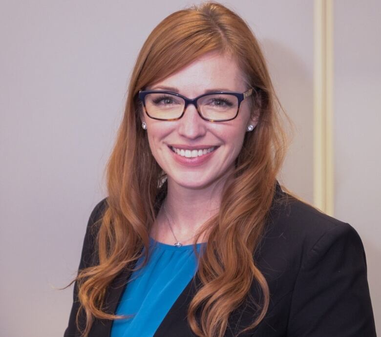 A woman in a black suit with blue shirt and wearing glasses.