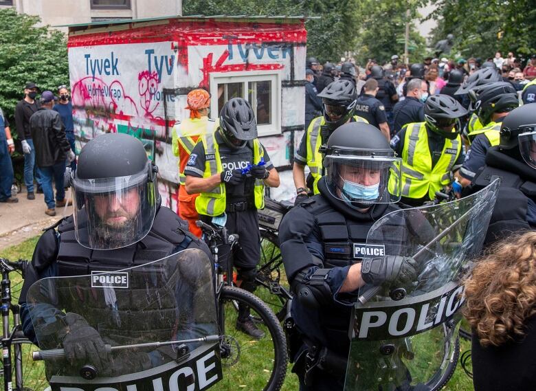 A group of police in riot gear including shields and batons form a circle around other police on bicycles and a man in construction clothes next to a wooden shelter. A large crowd is visible in the background