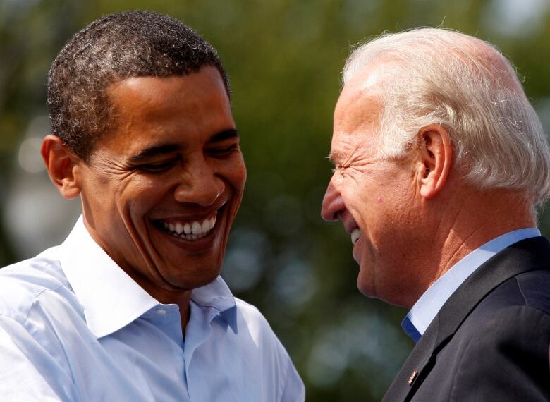 Two men, both smiling warmly, stand close to each other, face to face.