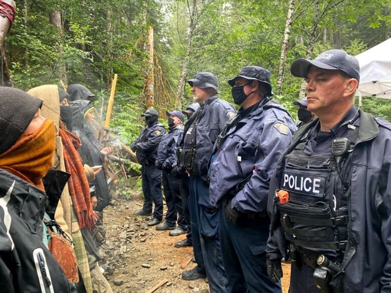 A row of protesters stands face-to-face with a row of police officers in a forest. 