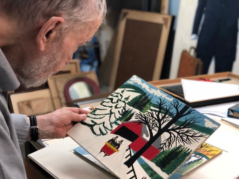 An older man with white hair and a beard looks at an authentic Maud Lewis painting.