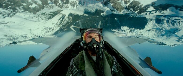 A man in pilot's gear sits in a cockpit surrounded by snowy mountains.