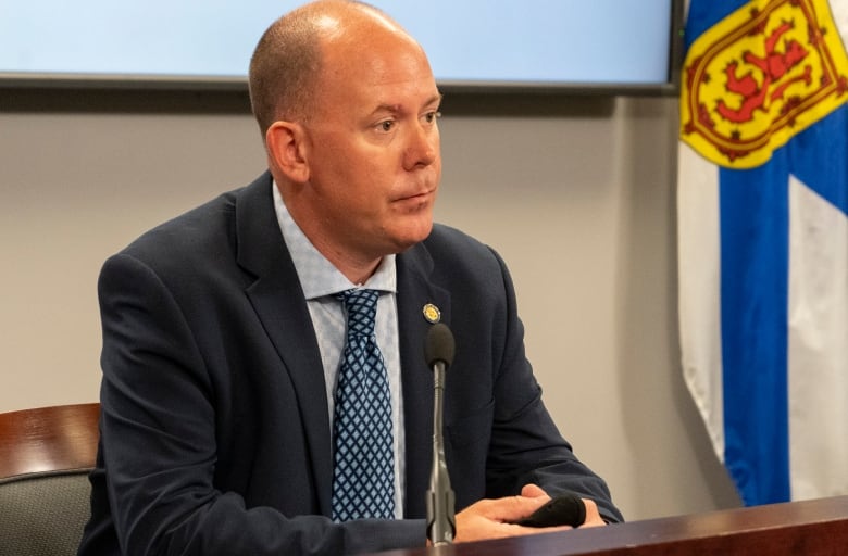 A bald man in a suit and tie sits at a desk with Nova Scotia flags behind him.