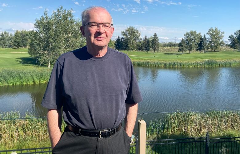 A white-haired man in glasses stands in front of a lake on a sunny day.