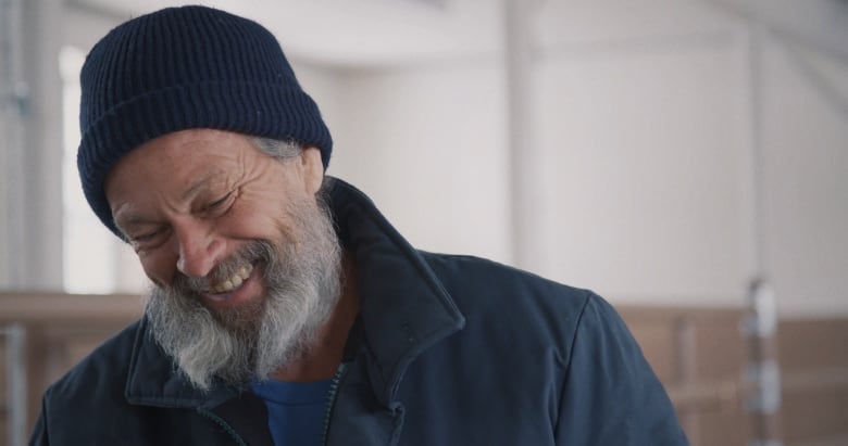 A man with grey hair and a grey beard is wearing a blue toque and smiling.