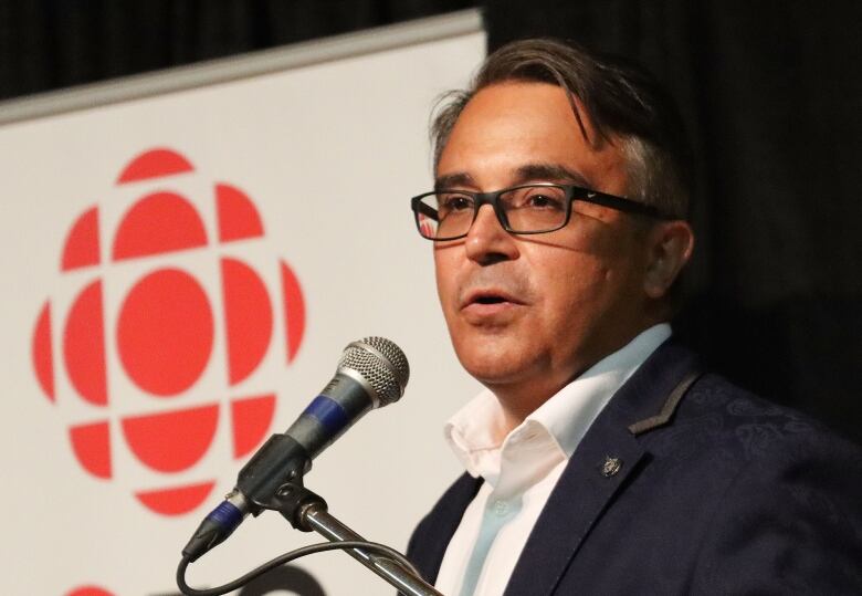 A man in a blue suit and glasses speaks into a microphone.