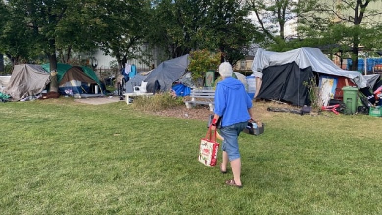 A person carrying a kit of some kind and a shopping bag walks toward a park encampment