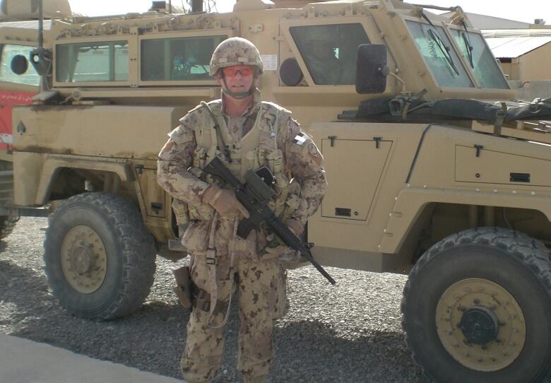 A man in Canadian military uniform holding a rifle stands by an amoured military military vehicle in Afghanistan.