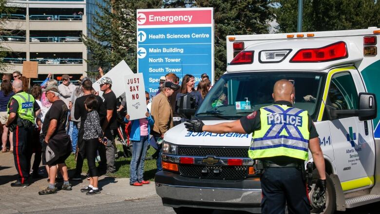 Police direct an ambulance past a crowd of people, one of whom is carrying a sign that says Just say no.