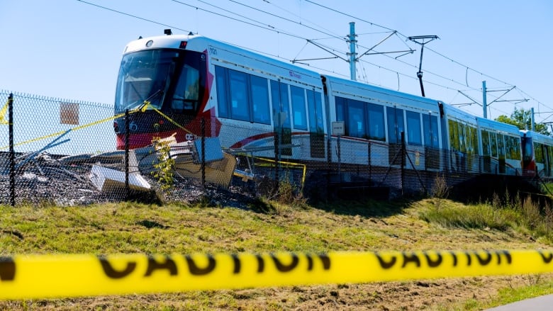 A red-and-white train is parked on train tracks on a sunny day while yellow tape with the word 