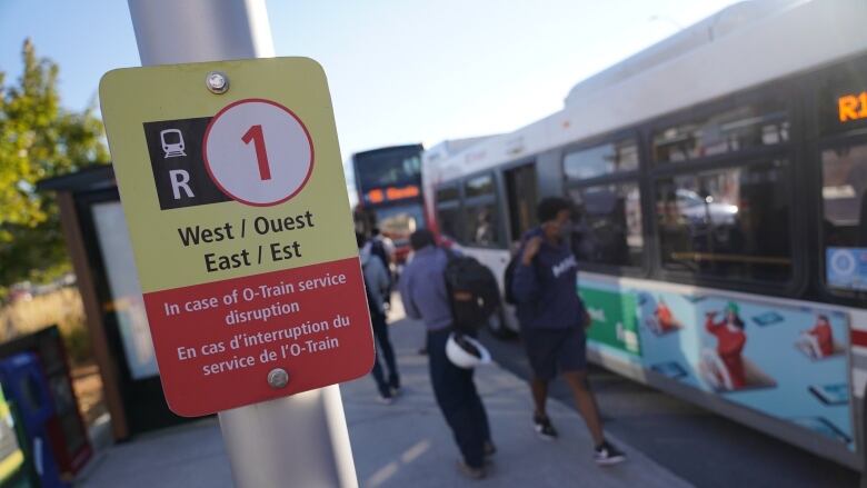 A yellow and red sign describes replacement bus service. In the background people walk by a bus.