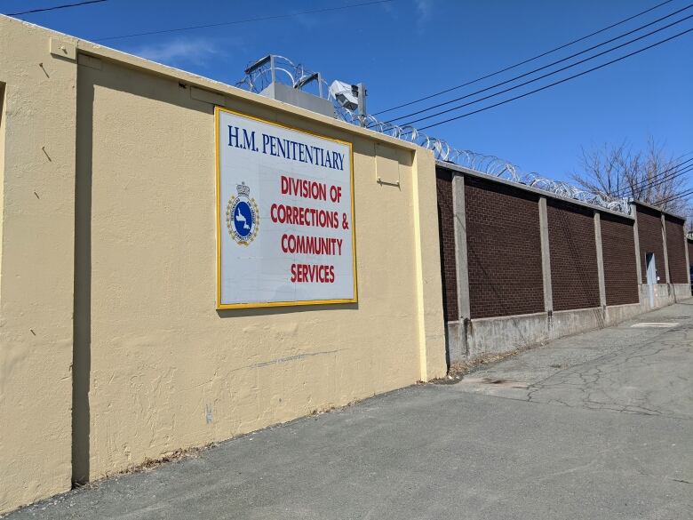 The front entrance of Her Majesty's Penitentiary in St. John's, NL. The prison sign is hung on a pale yellow wall, bordering a brick wall with barbed wire on top.