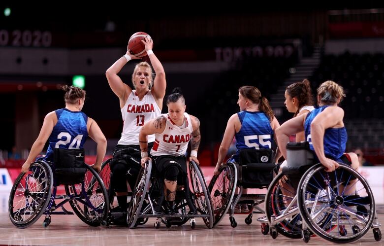 A blond woman in a wheelchair wearing a #13 Team Canada white jersey with red lettering is surrounded by teammates and opponents in wheelchairs as she shoots for the basket.