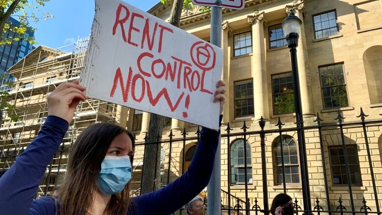 A woman in a surgical mask holds up a sign that says in red lettering, rent control now.