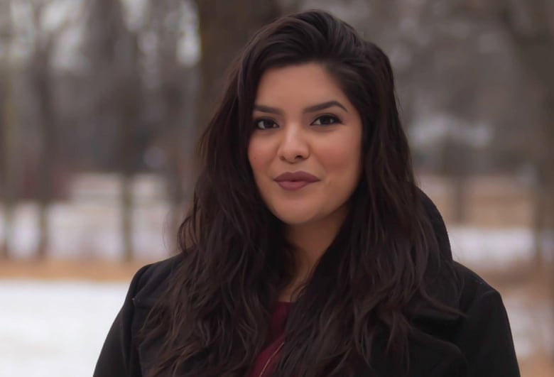 A woman in a long black coat stands outside in a treed and partially snow-covered area.