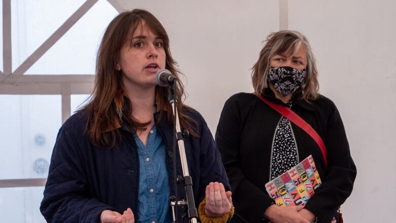 A woman stands speaking at a microphone as another woman in a mask looks on.