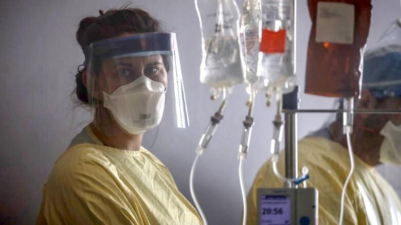 A health care worker in a mask and face shield looks past the camera while care for a patient in the ICU.