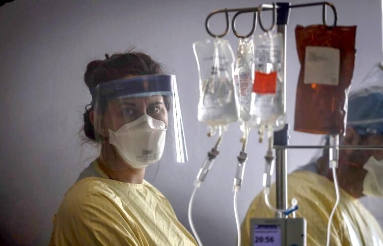 A health care worker in a mask and face shield looks past the camera while care for a patient in the ICU.