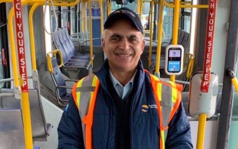 A man in a safety vest and ball cap stands in the middle of a city bus, smiling at the camera 