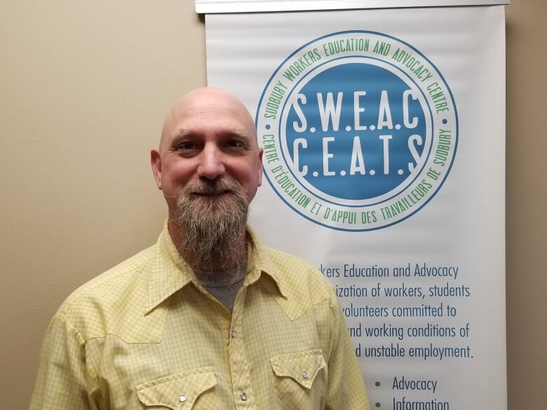 Bald man with goatee wearing a yellow shirt stands in front of a sign.