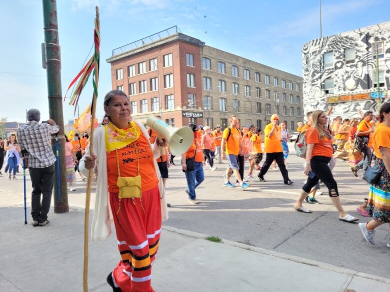 A woman in an orange shirt and red ribbon skirt holds a megaphone at a parade.