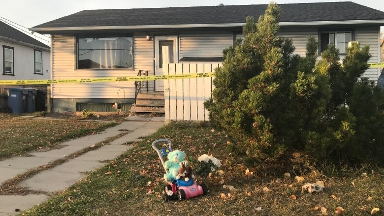 A small memorial consisting of flowers and toys appears on the front lawn of a bungalow with its entranceway blocked off by yellow crime-scene tape. 