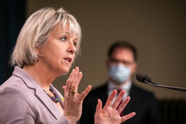 A white woman gestures at a podium while a masked man stands in the background.