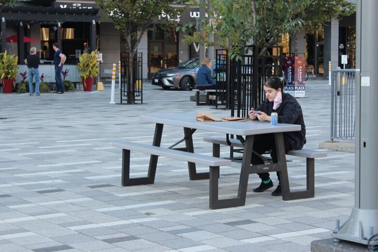 A woman with a face mask hanging off of her ear sits at a park bench and watches her cell phone