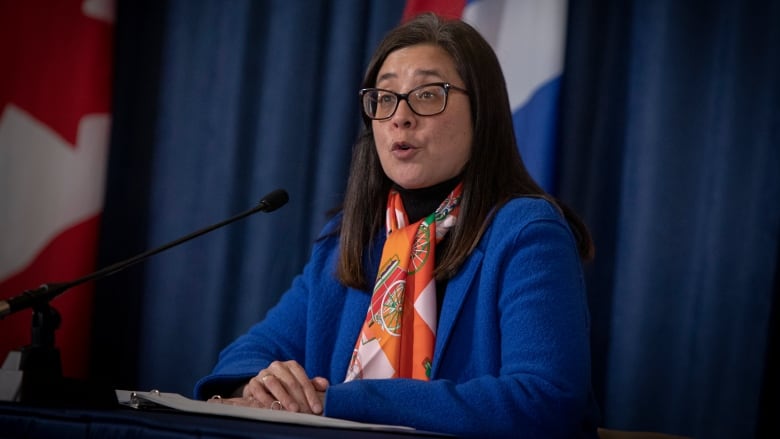 A woman wearing glasses speaks at a desk 