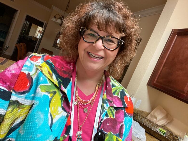 A woman with glasses wears a brightly coloured top and smiles at the camera.