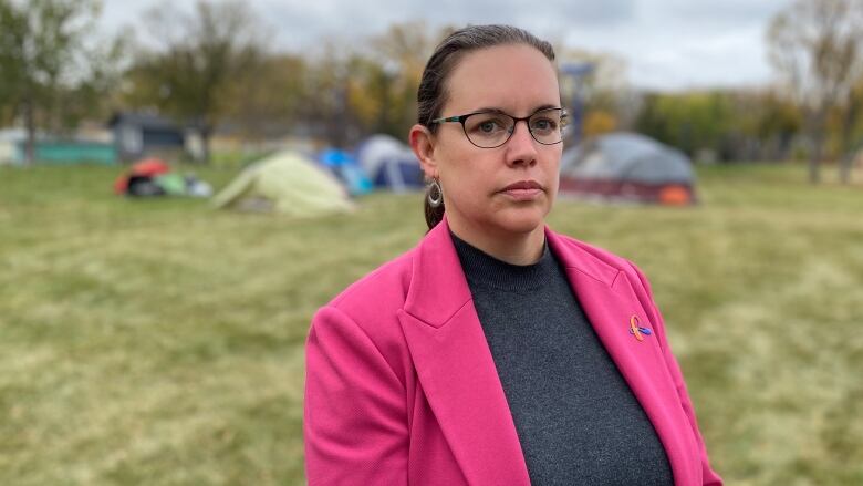 A woman in a pink blazer stand in a field where a homeless tent city stands. 